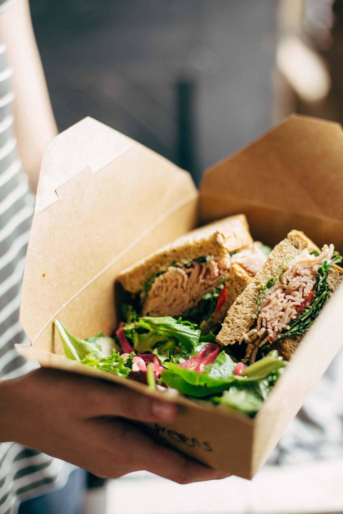 Hands holding a box lunch with a salad and a sandwich.